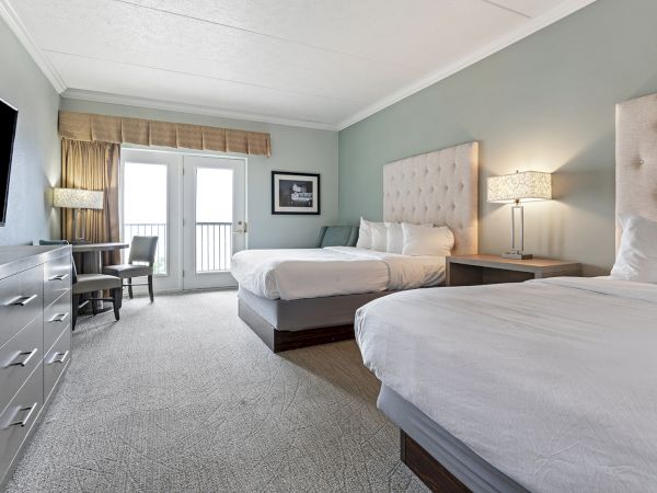 A hotel room with two beds, a TV, a dresser, a desk, a lamp on a nightstand, and a balcony with a view.