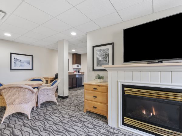 The image shows a cozy living area with a TV above a fireplace, wicker chairs around a table, and a small kitchen area in the background.