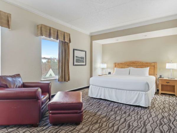 The image shows a spacious hotel room with a king-sized bed, armchair, ottoman, nightstands, and two windows with curtains.