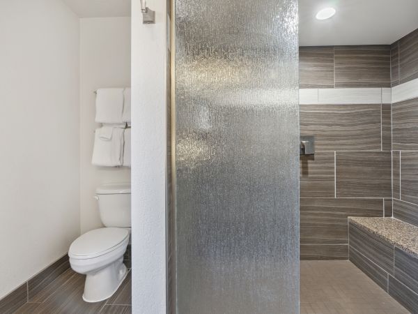 This image shows a modern bathroom with a toilet, towel rack, and a glass-enclosed shower area with grey tiles and a built-in bench.