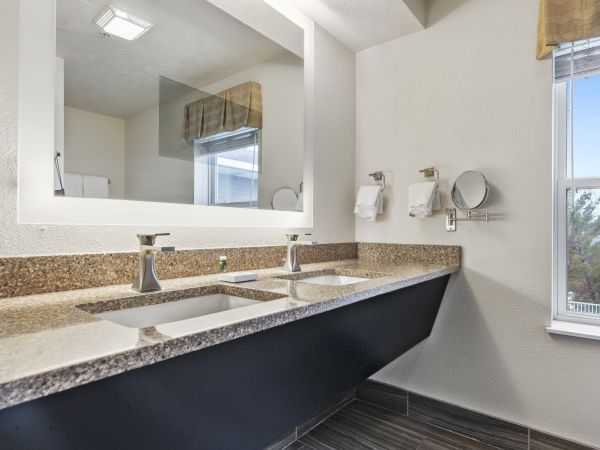 The image shows a modern bathroom with a large mirror, double sinks with sleek faucets, towel holders, and a window allowing natural light.