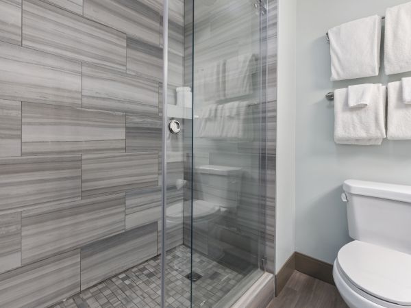 The image shows a modern bathroom with a glass-enclosed shower, gray tile walls, a white toilet, and white towels hung on the wall.