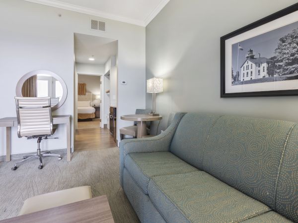 This image shows a living area with a green couch, a photograph on the wall, a desk with a chair, a small dining table, and a visible bedroom.