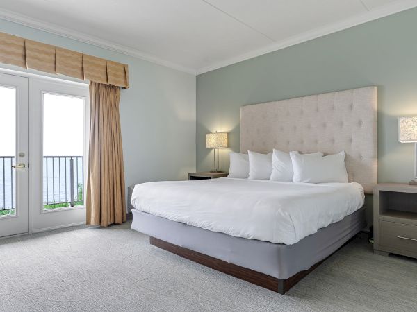 The image shows a modern bedroom featuring a large bed with a tufted headboard, two bedside tables with lamps, and French doors that open onto a balcony.