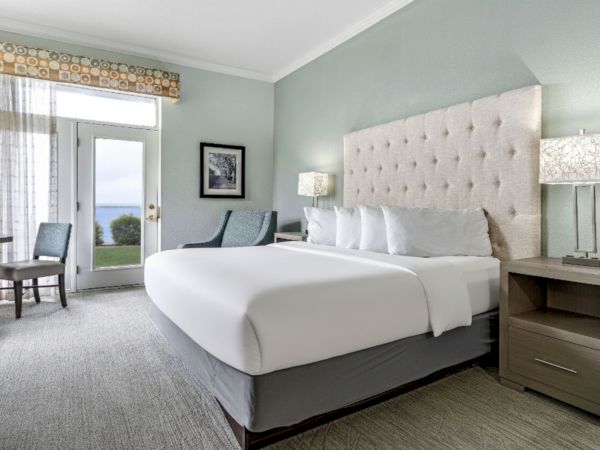 The image shows a well-lit hotel room with a large bed, lamps on nightstands, chairs, a table, a window, and a door leading outside.