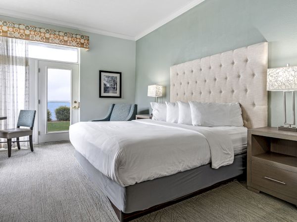 The image shows a well-lit hotel room with a large bed, lamps on nightstands, chairs, a table, a window, and a door leading outside.