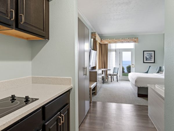 A hotel room with a kitchenette, bed, desk, TV, and a view of the balcony with chairs through a large window.