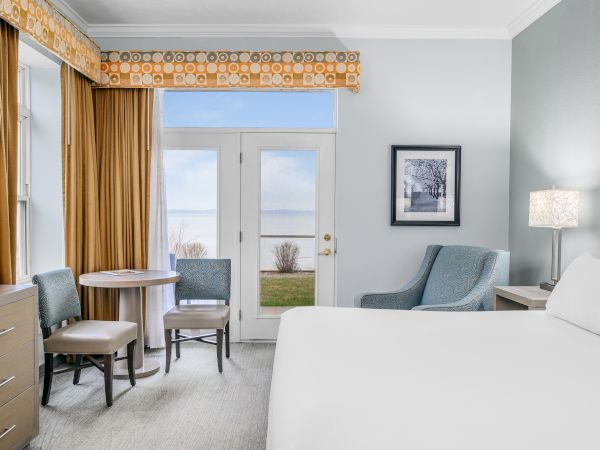 A cozy hotel room with a bed, armchair, small table and chairs, dresser, lamp, and French doors opening to an outside view, ending the sentence.