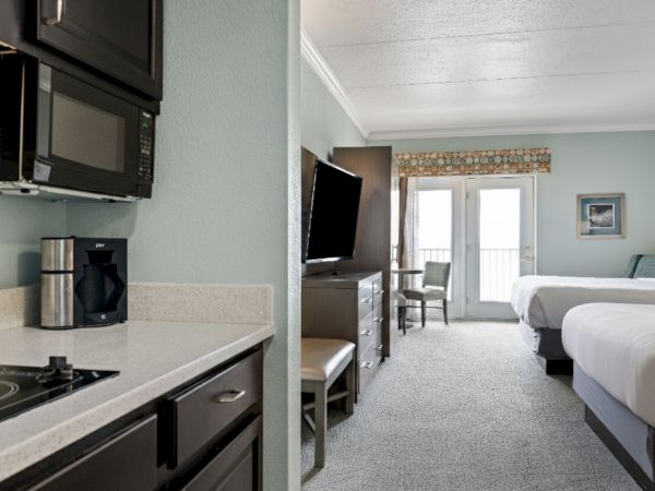 A hotel room with a kitchenette, TV, two beds, and a balcony door. The kitchenette has a stove, microwave, and coffee maker.