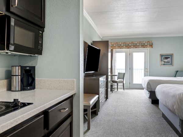 A hotel room with a kitchenette, TV, two beds, and a balcony door. The kitchenette has a stove, microwave, and coffee maker.