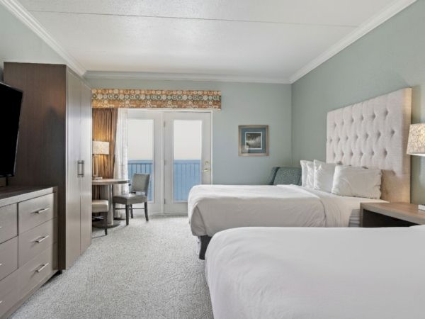 A hotel room with two double beds, a large TV on a dresser, a small table with chairs, and a window or door leading to a balcony; bedside lamp is on.