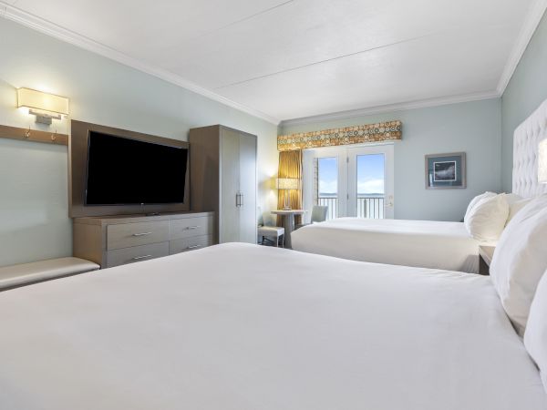 This image shows a hotel room with two large beds, a wall-mounted TV, a wardrobe, a bench, a desk, and a balcony with a view outside.