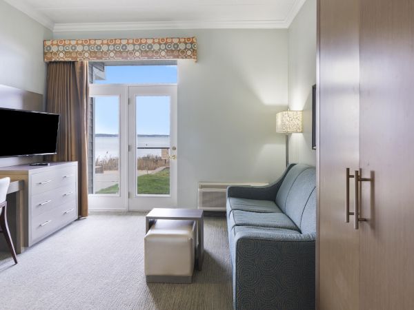 A room featuring a sofa, TV, desk, and chair with a view of a body of water through a french door, and a lamp on the wall.