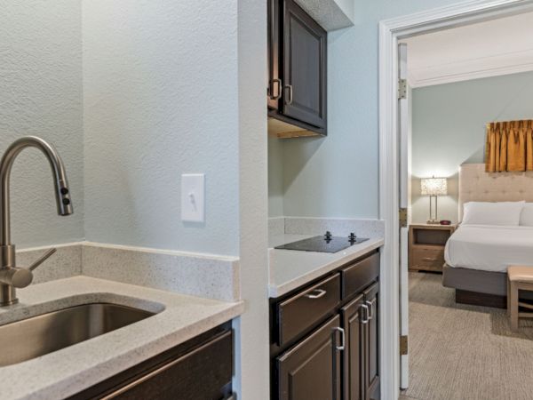 The image shows a kitchenette with a sink and stove, leading into a bedroom with a neatly made bed and a bedside table.