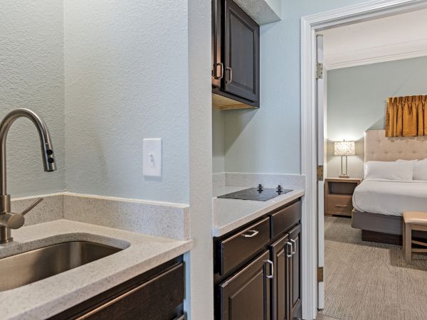 The image shows a kitchenette with a sink and stove, leading into a bedroom with a neatly made bed and a bedside table.