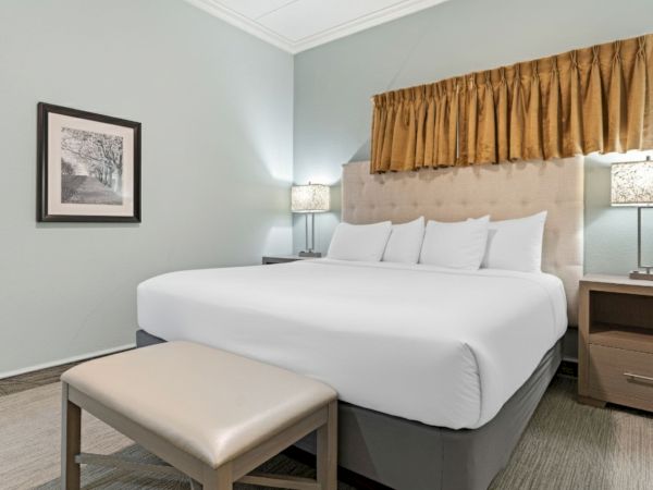 A neatly made bed with white linens, bedside tables with lamps, a framed picture, and a cushioned bench at the foot of the bed.