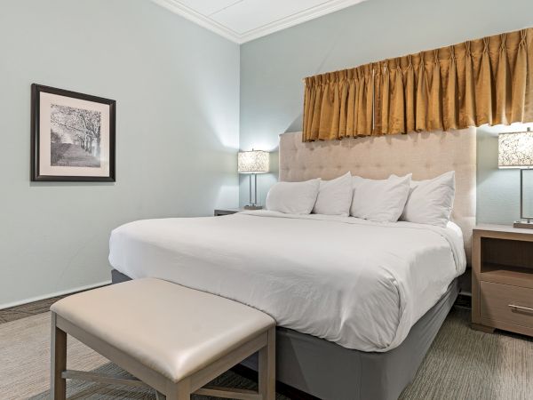 A neatly made bed with white linens, bedside tables with lamps, a framed picture, and a cushioned bench at the foot of the bed.