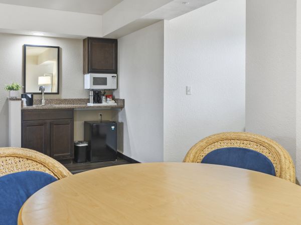 The image shows a small kitchenette with a microwave, mini fridge, and sink, next to a round wooden dining table with four wicker chairs.