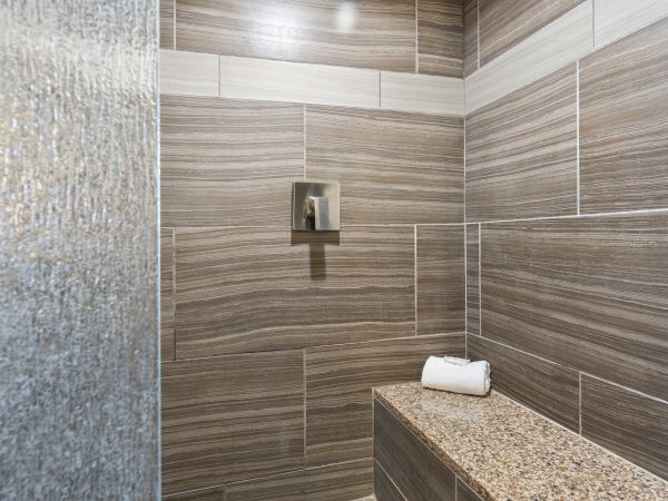 The image shows a modern bathroom shower area with brown and beige tiles, a metallic faucet, and a granite bench with a rolled white towel on top.