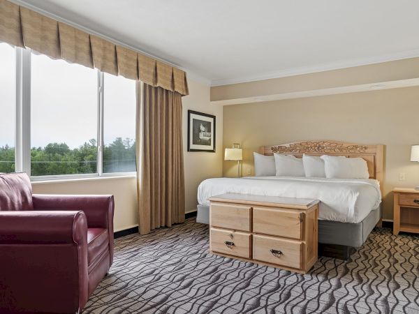 A hotel room with a large bed, bedside tables with lamps, an armchair, a wooden chest, and a window with drapes and a nature view, ending the sentence.