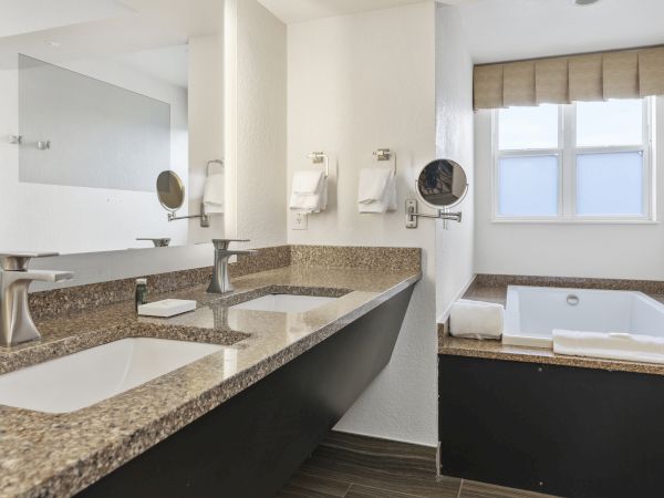 A modern bathroom with double sinks, a large mirror, wall-mounted towels, and a bathtub next to a window, featuring a minimalist design.
