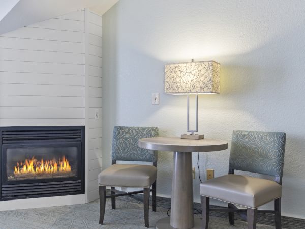 A cozy living area with a fireplace, a round table, two chairs, and a lamp on the table against a light-colored wall.