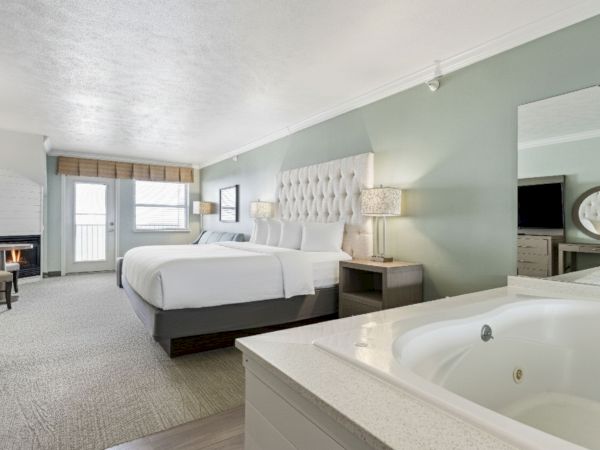 A modern hotel room featuring two beds, a hot tub, a large mirror, a desk, and a TV. The room has soft lighting and a view through French doors.