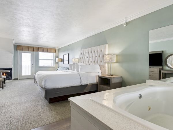 A modern hotel room featuring two beds, a hot tub, a large mirror, a desk, and a TV. The room has soft lighting and a view through French doors.