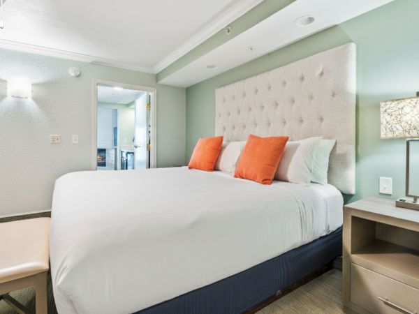 A modern bedroom with a large bed, white linens, orange pillows, a cushioned headboard, a bench, and a nightstand with a lamp.