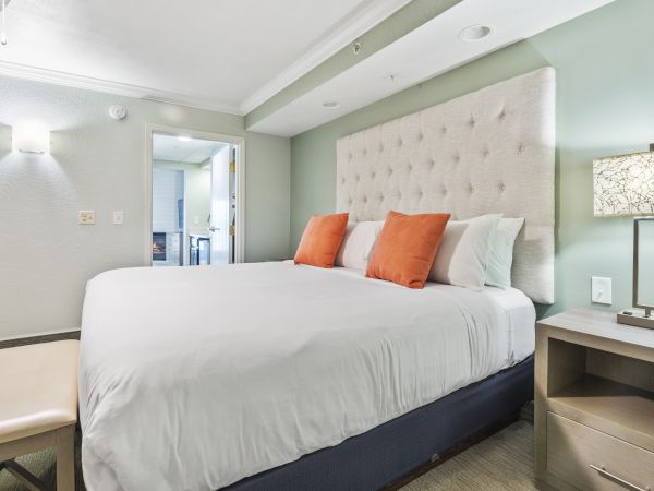 A modern bedroom with a large bed, white linens, orange pillows, a cushioned headboard, a bench, and a nightstand with a lamp.