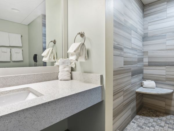 A modern bathroom featuring a sink with a silver faucet and towel, a mirror, and a tiled shower with a bench and white towels.
