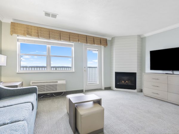 A cozy living room with a sofa, ottoman, TV on a dresser, fireplace, window, and door leading to a balcony with a scenic view of blue sky and clouds.