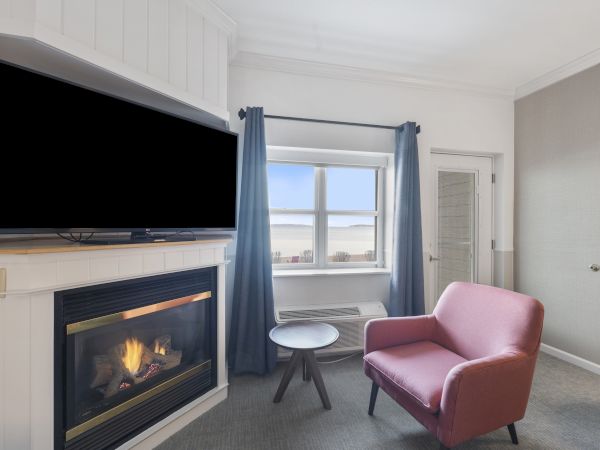 A cozy room with a fireplace, TV, pink chair, small table, and a window with blue curtains overlooking an outdoor view.