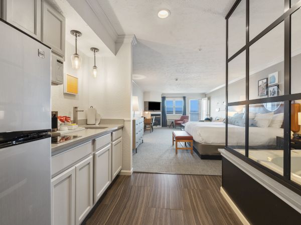 The image shows a spacious hotel room featuring a kitchenette, a large bed, modern furnishings, and a sitting area with windows in the background.