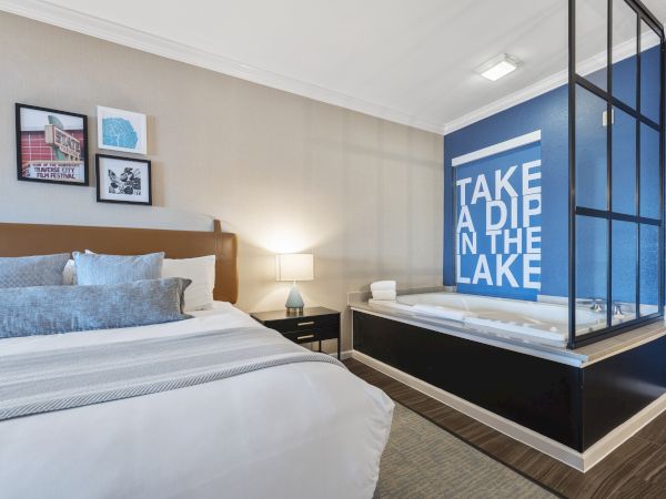 A modern bedroom with a comfortable bed, wall art, a lamp on a nightstand, and an in-room bath area with a blue wall reading "TAKE A DIP IN THE LAKE."