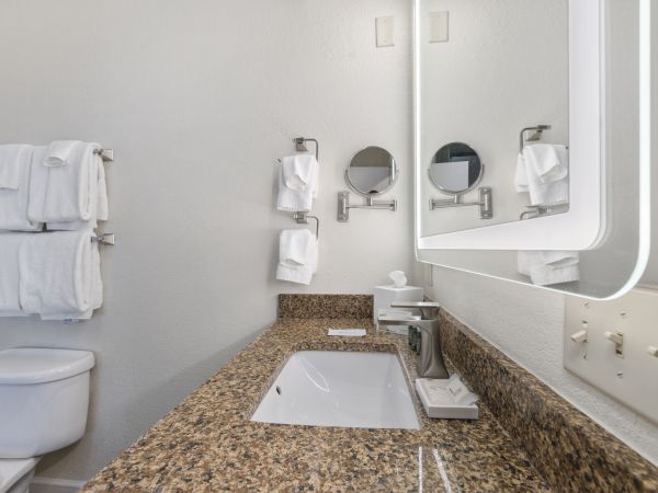 The image shows a modern bathroom with a granite countertop, sink, wall-mounted mirrors, towels, and a lit mirror on the wall.