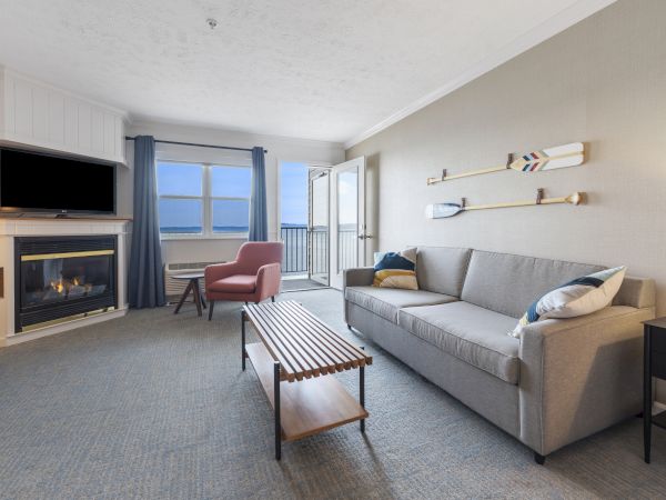 A living room with a sofa, coffee table, fireplace with TV, armchair, balcony with ocean view, and decorative oars on the wall.