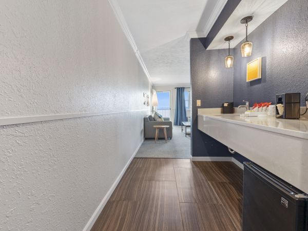 The image shows a hotel room with a hallway leading to a seating area. There’s a mini fridge, countertop, and lights in the foreground.
