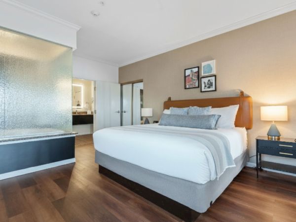 A modern bedroom with a large bed, a side table with a lamp, decor on the wall, and an open bathroom area with a glass shower and a wooden floor.