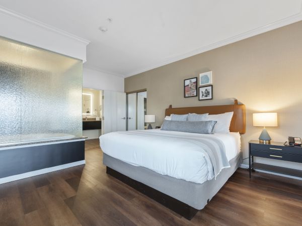 A modern bedroom with a large bed, a side table with a lamp, decor on the wall, and an open bathroom area with a glass shower and a wooden floor.