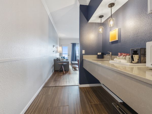 A hallway with a kitchenette on the right, leading to a living area with a sofa, coffee table, and curtains drawn to one side at the window.