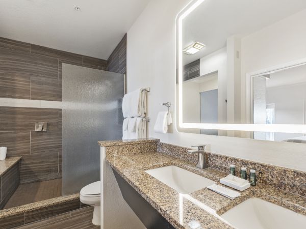 The image shows a modern bathroom with a double sink vanity, granite countertops, a large mirror with lighting, towels, and a walk-in shower.