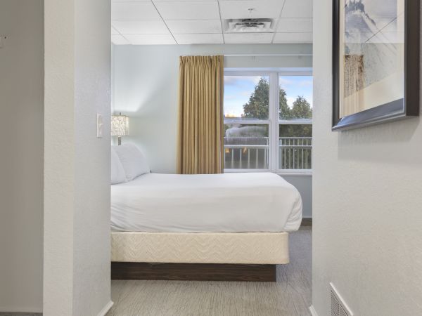 The image shows a modern hotel room with a neatly made bed, a window with yellow curtains, a bedside lamp, and artwork on the wall.