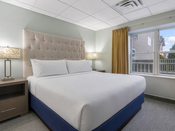 A clean hotel room with a king bed, beige headboard, two lamps, and a large window overlooking a balcony, all in soothing neutrals.