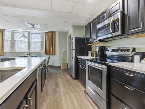 A modern kitchen with stainless steel appliances, dark cabinets, a microwave, a coffee maker, and a dining area with windows and curtains.