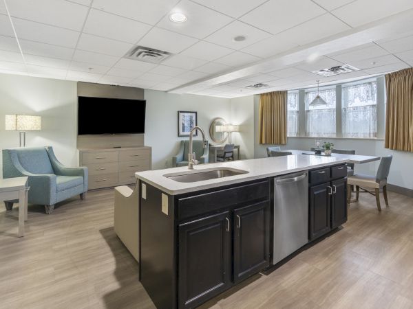 A modern room with a kitchen island and sink, comfortable seating area, flat-screen TV, and a dining table with chairs, large windows with curtains ending the sentence.