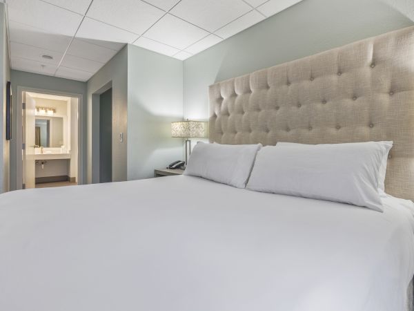 The image shows a neatly made bed with white linens in a modern hotel room, featuring a tufted headboard and a view leading into a bathroom.