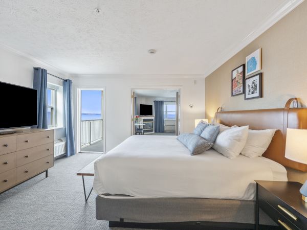 A bright bedroom features a large bed, dresser, TV, balcony with ocean view, and modern decor on the walls, creating a cozy atmosphere.