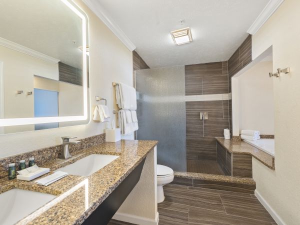 A modern bathroom features a double sink vanity, backlit mirror, stacked towels, a toilet, a walk-in shower with frosted glass, and a large bathtub.
