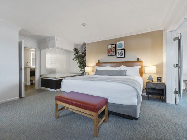 This image shows a hotel room with a large bed, two bedside tables with lamps, a bench at the foot of the bed, and an adjacent bathroom.